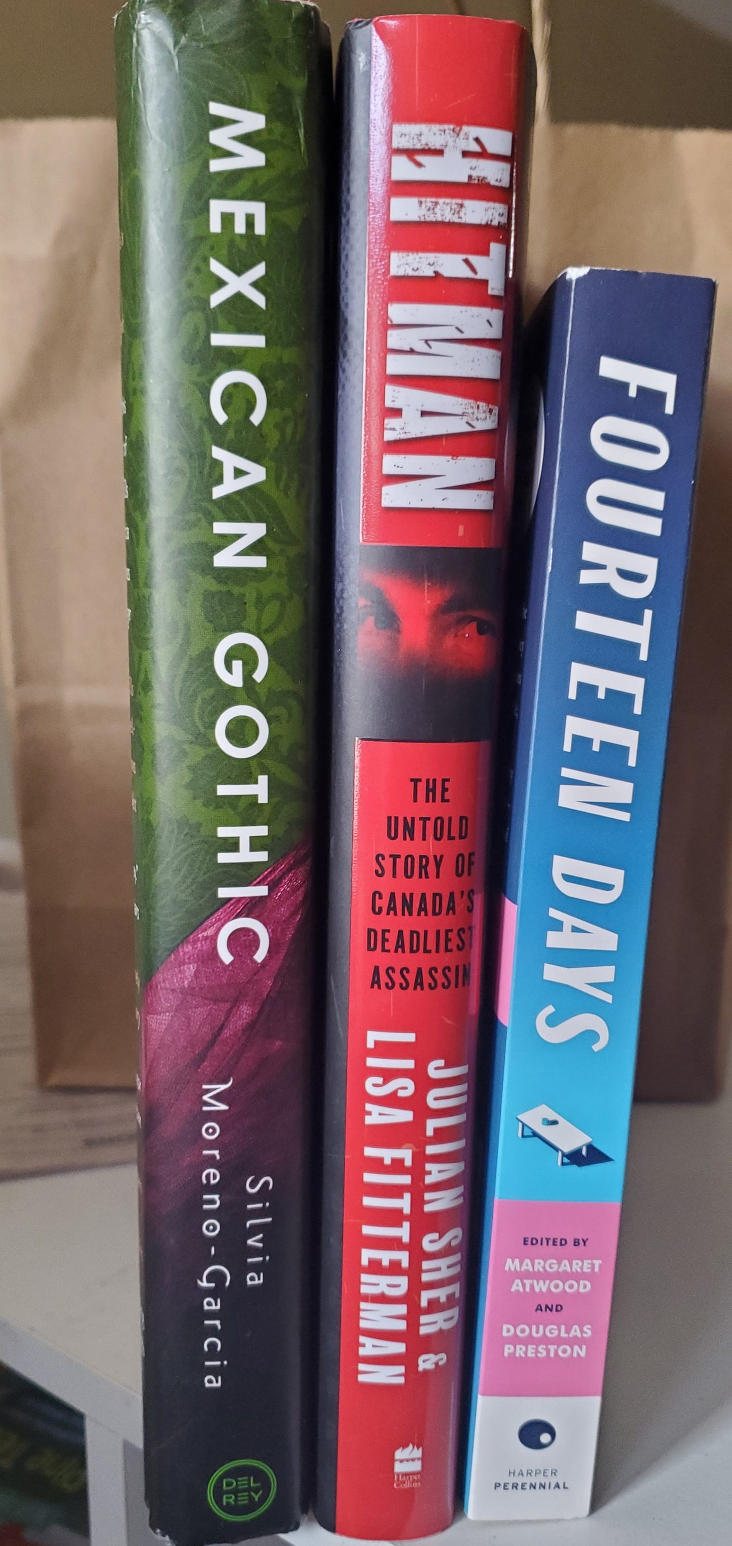 Three books standing with their spines out: Mexican Gothic by Sylvia Moreno-Garcia, Hitman by Julina Sher and Lisa Fittermna, & Fourteen Days by Margaret Atwood and Douglas Preston.