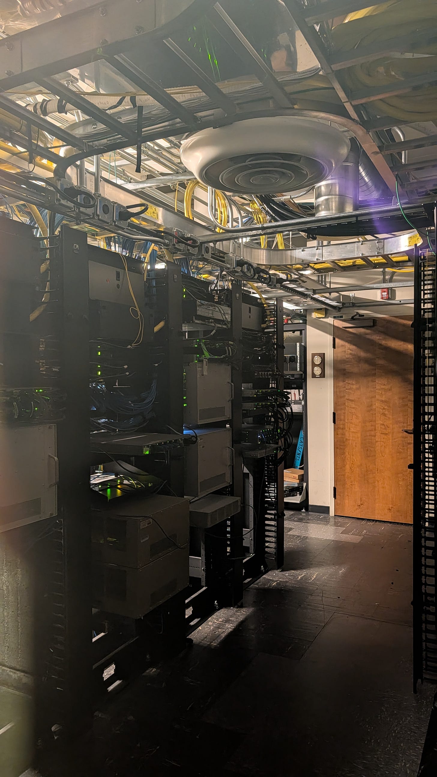 Server and networking racks with green indicator lights, seen through the window of a locked door in the Communications Building basement.