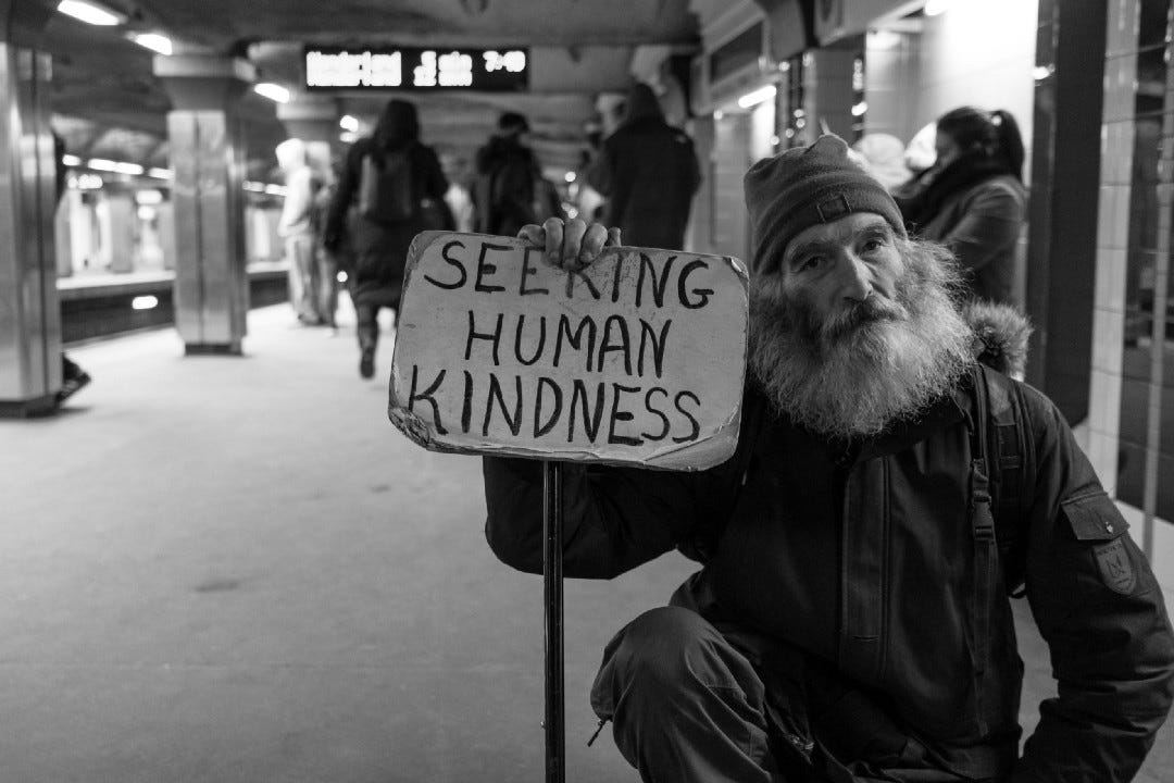 A bearded homeless man holds up sign saying "seeking human kindness"