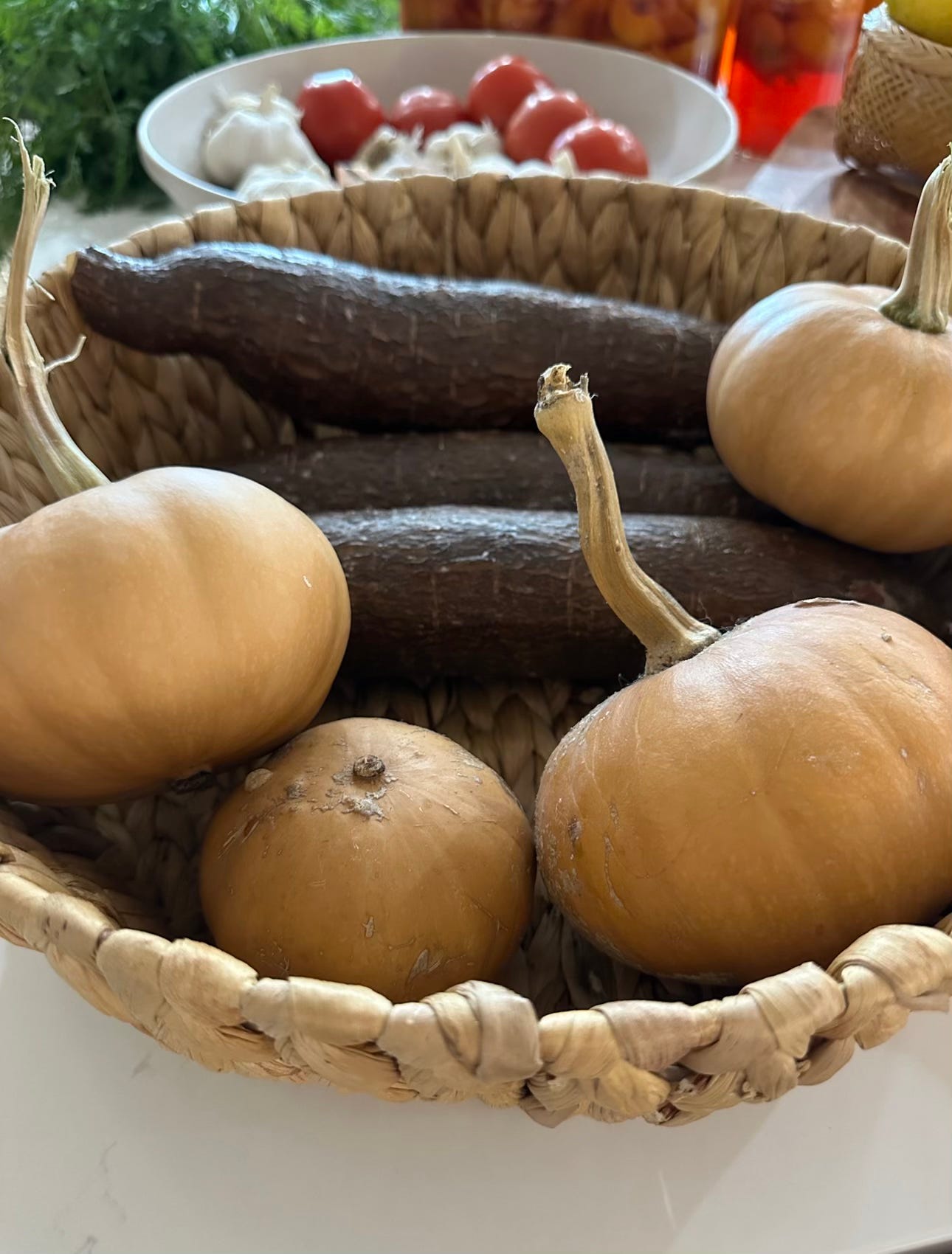 Woven basket with four tan pumpkins and two brown cassava roots. A bowl of garlic and tomatoes in the background.