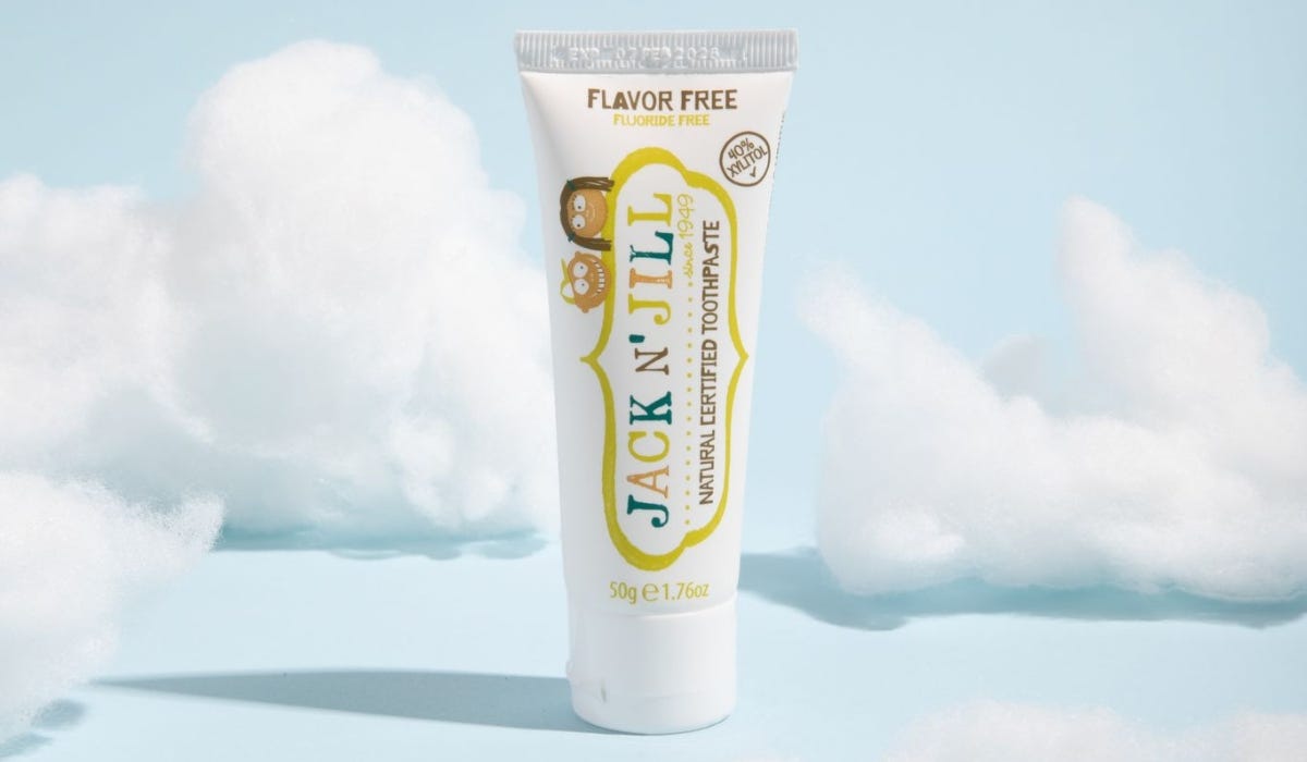 A tube of Jack N' Jill natural certified toothpaste, flavor and fluoride-free, against a light blue background with fluffy clouds.