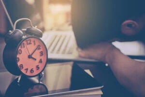 Close up of a man resting head on his desk with an alarm clock in the foreground