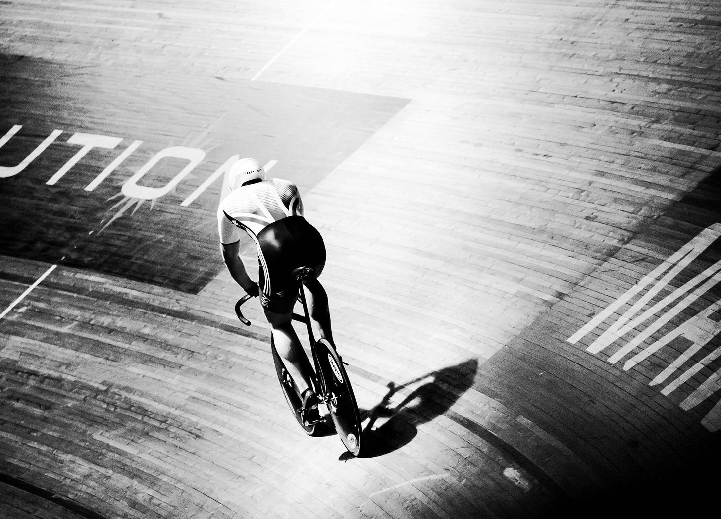 A lone track cyclist rides through a brightly lit velodrome, casting a long shadow across the wooden track. A lone track cyclist rides through a brightly lit velodrome, casting a long shadow across the wooden track.