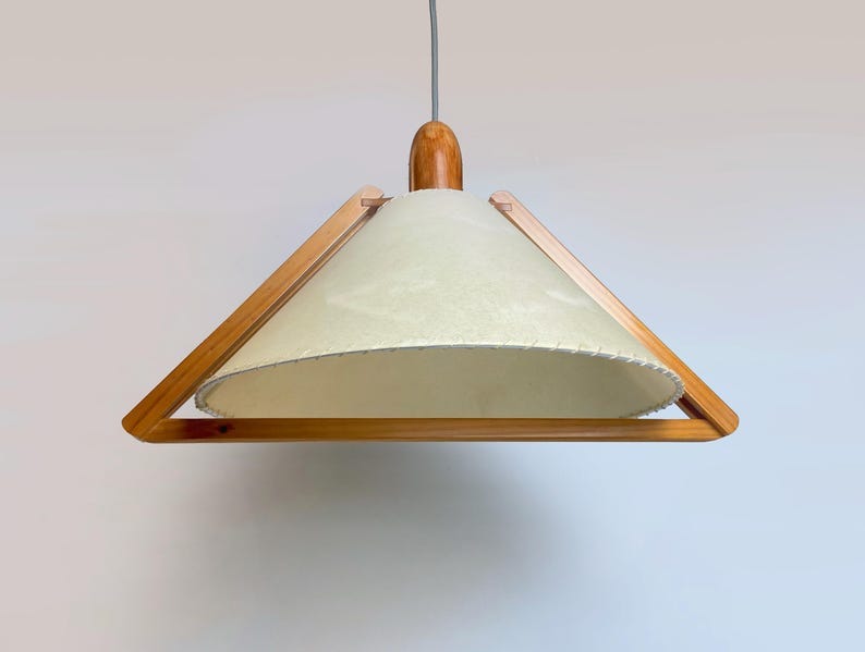 May include: A mid-century modern pendant light with a light cream-colored shade and a wooden frame. The shade has a textured appearance and is trimmed with a decorative edge. The light fixture is suspended from a gray cord and has a wooden top.