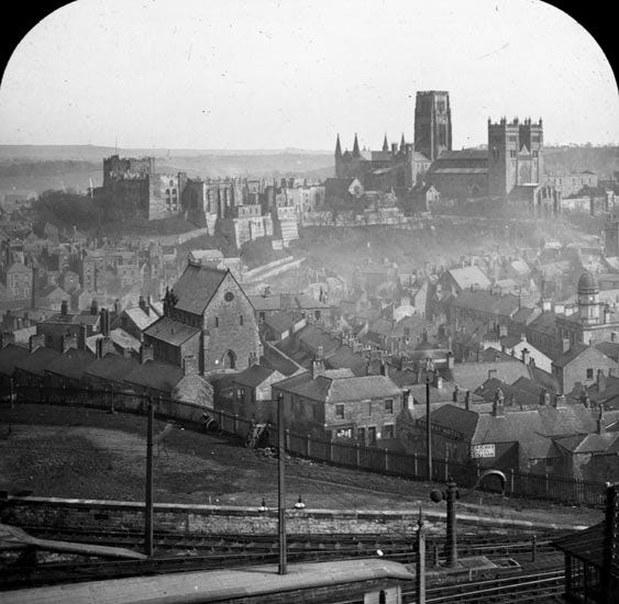 Black and white historical photograph of a hilly town skyline featuring various buildings, including churches and residential rooftops, with a distant view of ruins or a castle.