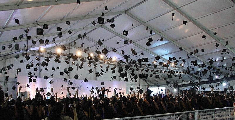 A large crowd of graduating students in gowns, seen from the back of the room, with the air overhead filled with mortar-board hats that have been tossed high and are likely to cause injury or incident on landing.