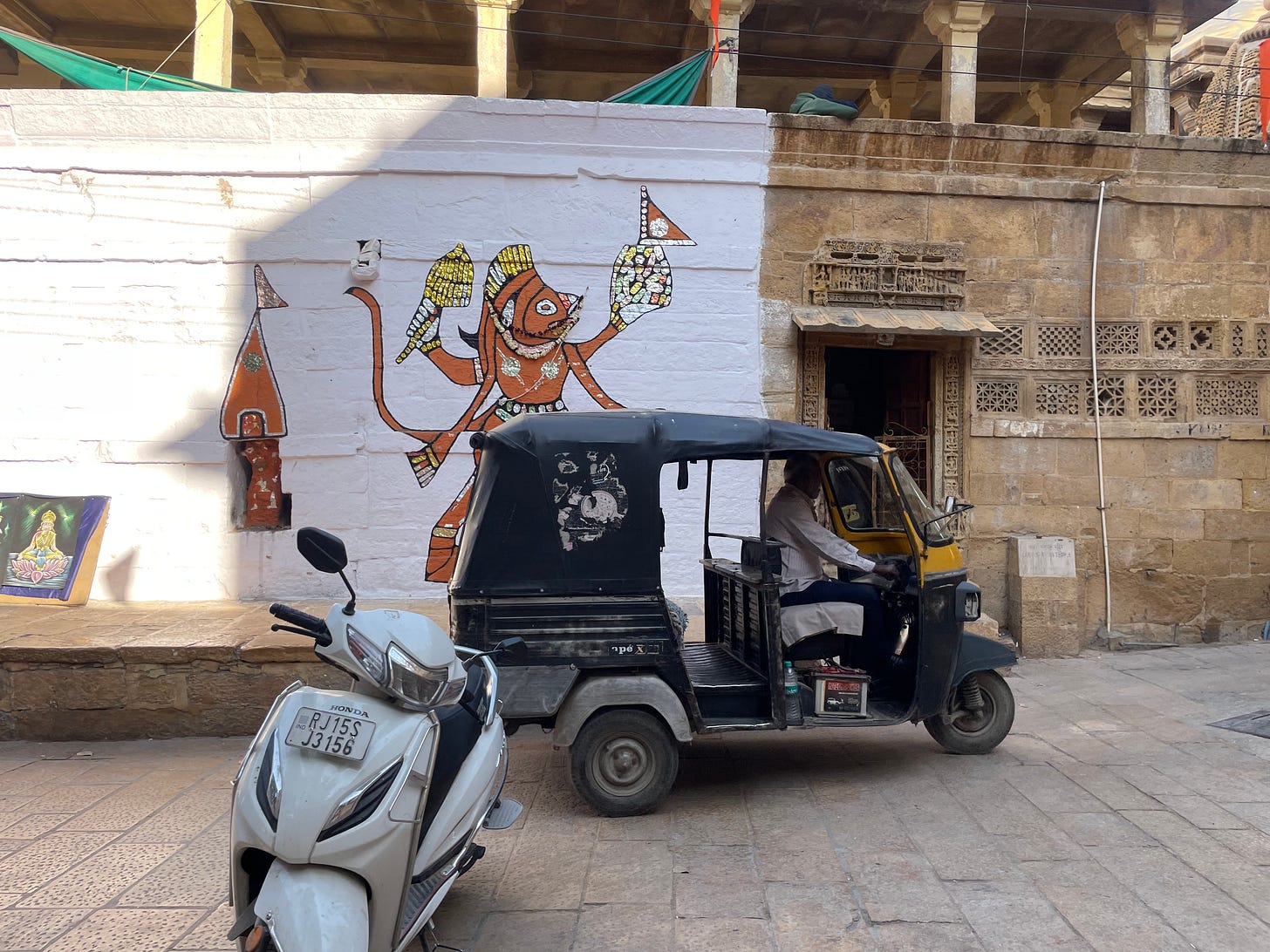 A driver sits in a rickshaw before a whitewashed wall painted with religious graffiti, and a sandstone wall with intricate carved windows. A driver sits in a rickshaw before a whitewashed wall painted with religious graffiti, and a sandstone wall with intricate carved windows.