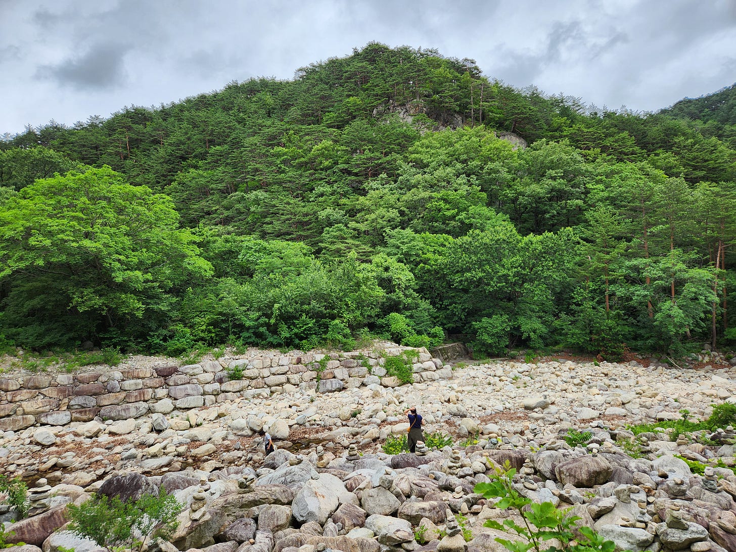 a tiny human figure (me) standing in a rockscape that looks like a dry riverbed looking at the background of mountain. I am standing with rocks all around me, many of which are stacked. I am surrounded by prayers.