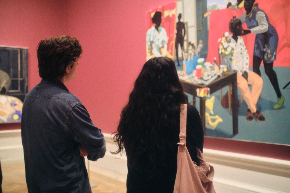 An exhibition view shows two visitors seen from behind looking at a large, brightly colored figurative painting on a pink gallery wall.
