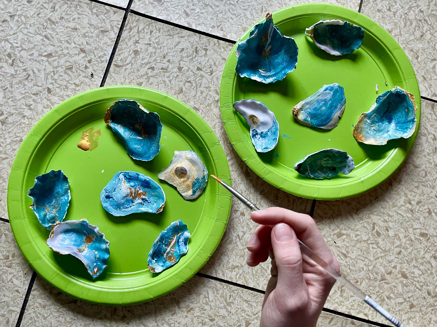 A photograph of two green plates, covered in hand-painted oyster shells. There is a hand holding a paint brush with some gold paint on the tip. A photograph of two green plates, covered in hand-painted oyster shells. There is a hand holding a paint brush with some gold paint on the tip.