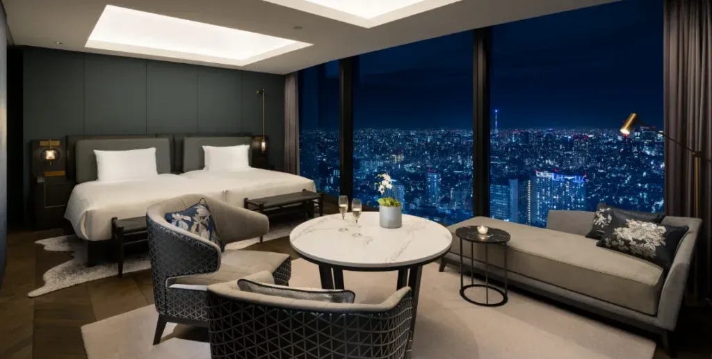 A luxurious hotel suite at night featuring twin beds with a gray upholstered headboard, a seating area with patterned armchairs and a sofa around a round marble table, and floor-to-ceiling windows showcasing a sweeping night view of the Tokyo skyline including Tokyo Tower in the distance. A luxurious hotel suite at night featuring twin beds with a gray upholstered headboard, a seating area with patterned armchairs and a sofa around a round marble table, and floor-to-ceiling windows showcasing a sweeping night view of the Tokyo skyline including Tokyo Tower in the distance.