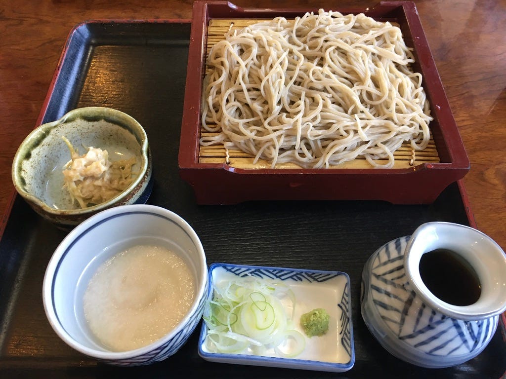 a tray of food with cold soba on zaru at the top, soy-based dipping sauce at bottom right, green and white scallions at bottom, white grated mountain yam at bottom left, and a vegetable side dish to the left a tray of food with cold soba on zaru at the top, soy-based dipping sauce at bottom right, green and white scallions at bottom, white grated mountain yam at bottom left, and a vegetable side dish to the left