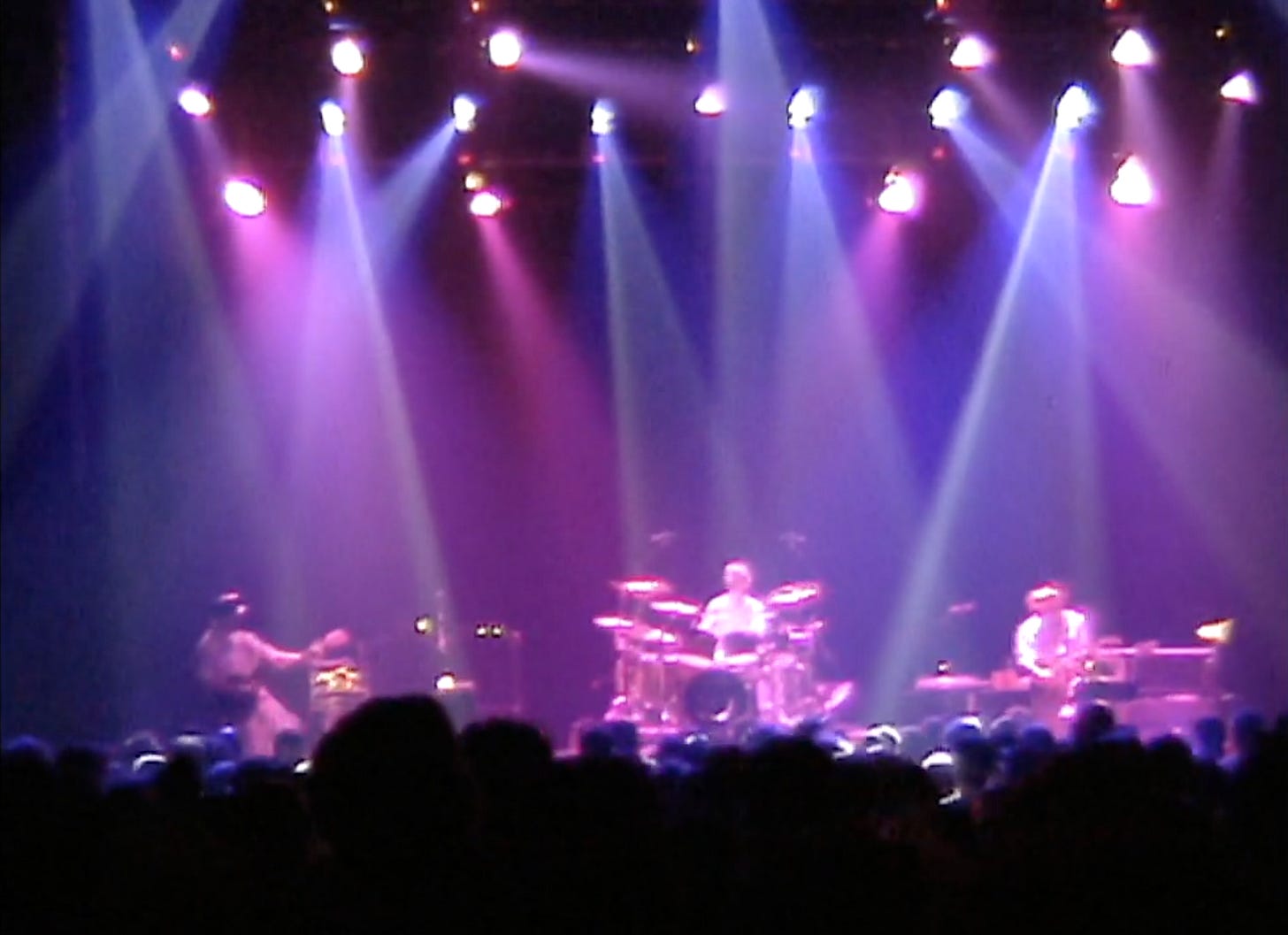 A videograb of the three musicians in Oysterhead performing on stage in purple light.