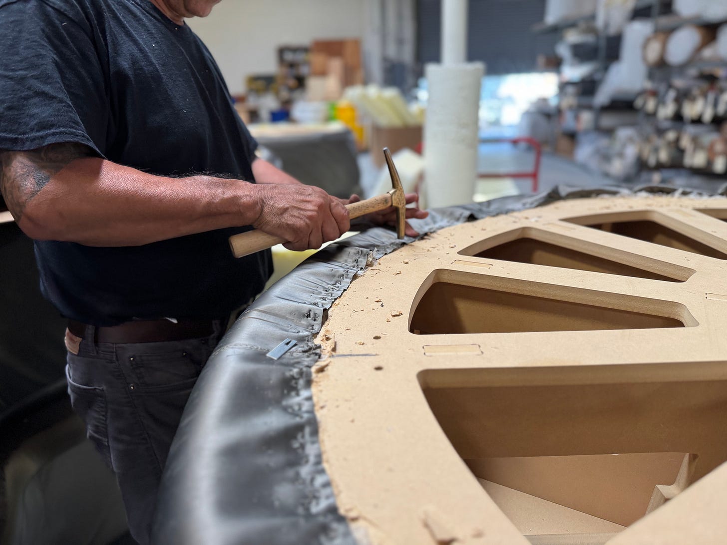 Close-up of an upholsterer using a hammer to secure black leather fabric over a large circular wooden frame.