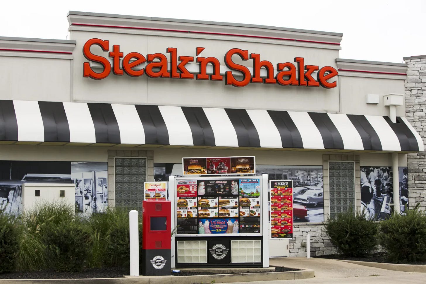 Exterior of a Steak 'n Shake restaurant featuring the red logo sign, black-and-white striped awning, and outdoor menu display board.