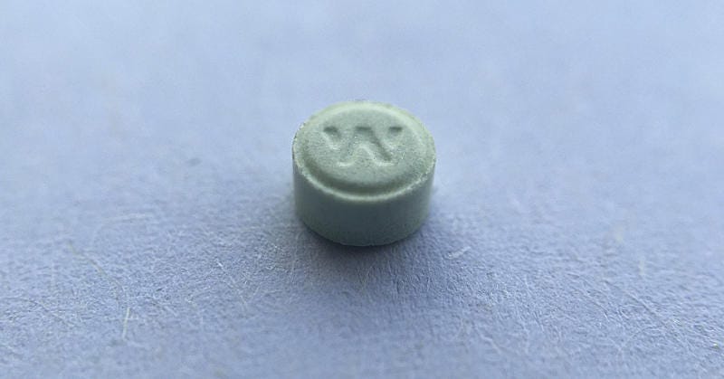 A small, round, pale green tablet photographed in close-up on a smooth blue-gray surface (paper), showing a debossed “W” on the top face.