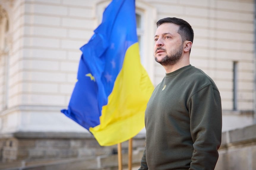 A man in army fatigues stands in front of the Ukrainian flag A man in army fatigues stands in front of the Ukrainian flag