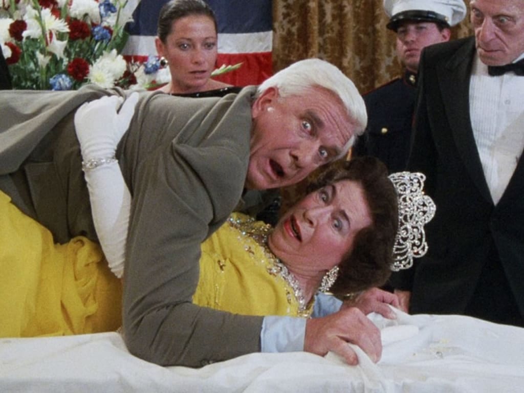 Frank Drebbin lying on top of queen elizabeth; they both look horrified as do bystanders.