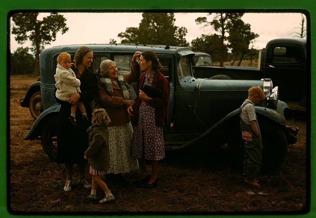 ALT TEXT: A color photograph of three smiling white women and three blonde children in 1940s clothing standing next to a dark green 1940s era automobile parked in a field, with several broadleaf trees in the background. One woman holds a baby, the next woman is an elder who gestures to the third woman's hair. The other two kids face away from the camera, thinking about whatever kids think about.