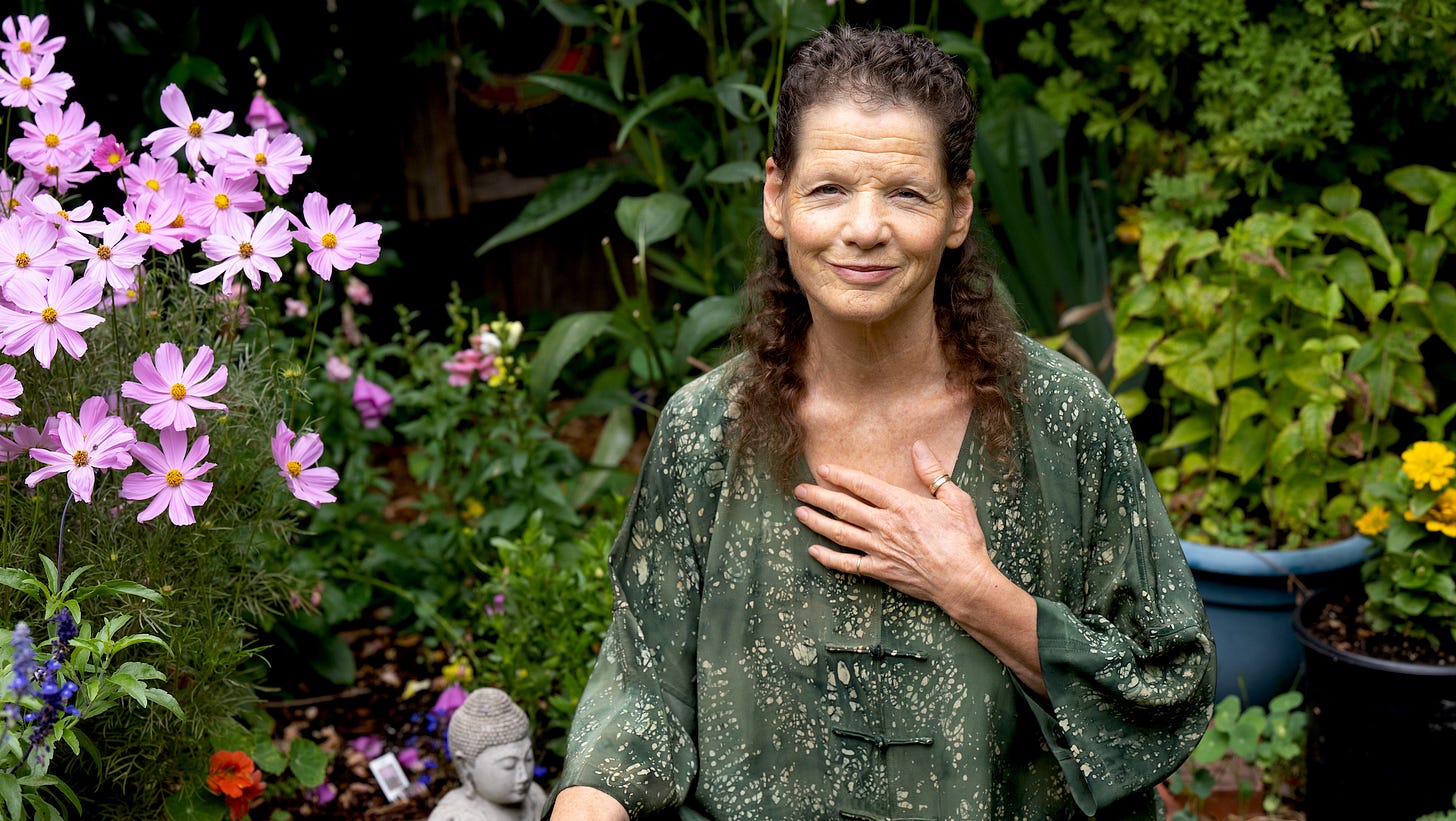 Liza surrounded by flowers and plants in an overgrown garden. Her hand is on her heart as she looks directly at the camera smiling gently.