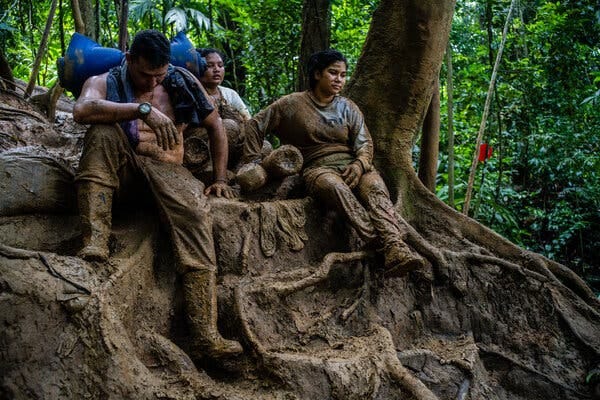 A small group of people sit in a forest on a steep, muddy path. They are covered in mud and look tired.