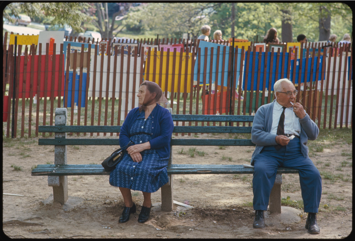 A woman in a blue patterned dress and headscarf sits on one end of a park bench while an elderly man in a light blue cardigan and dark pants sits on the opposite end, appearing to eat something. They sit with notable space between them. Behind them is a colorfully painted wooden fence with vertical slats in red, yellow, blue, and green. In the background, people can be seen walking in what appears to be a park or public space with trees.