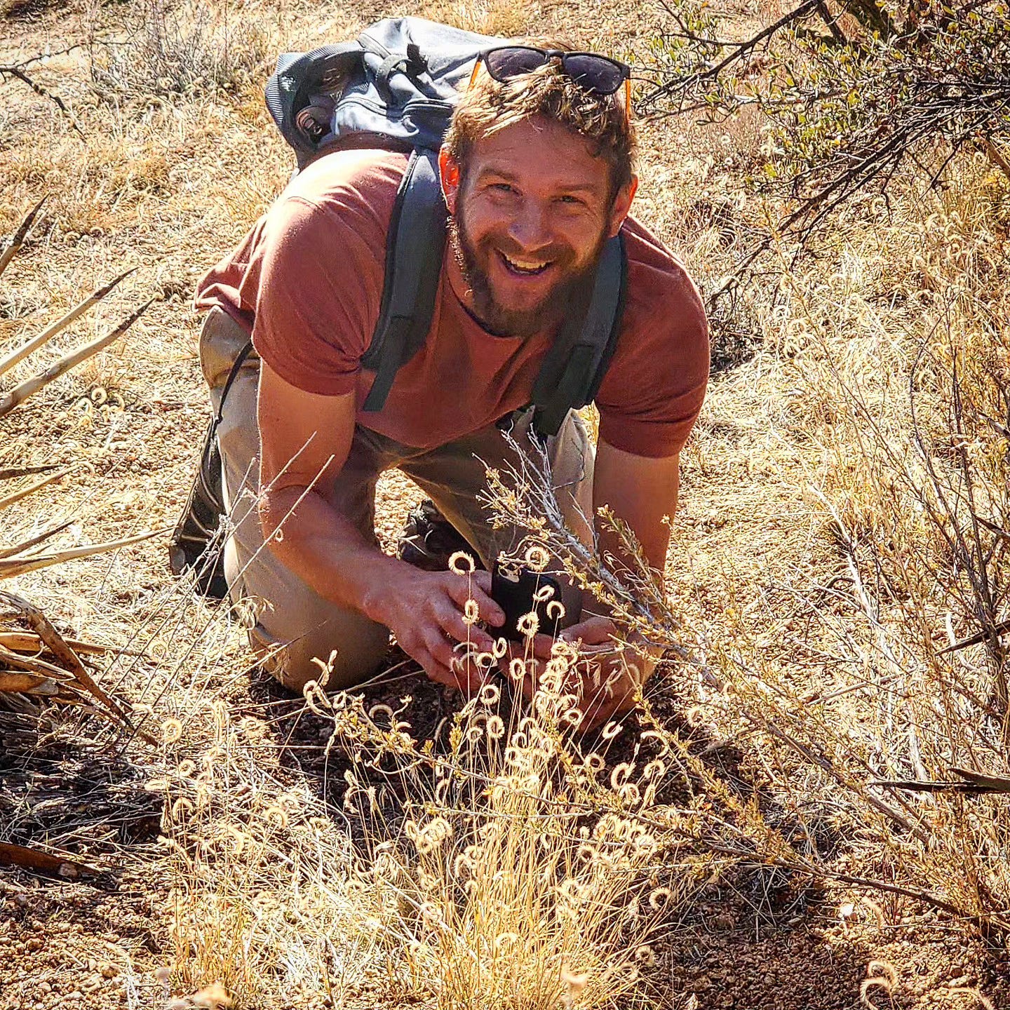 Photo of Stuart Soboleski, a grinning white man in short sleeves and hiking pants, kneeling down among some desert glasses. It's sunny out, and the light is radiant.
