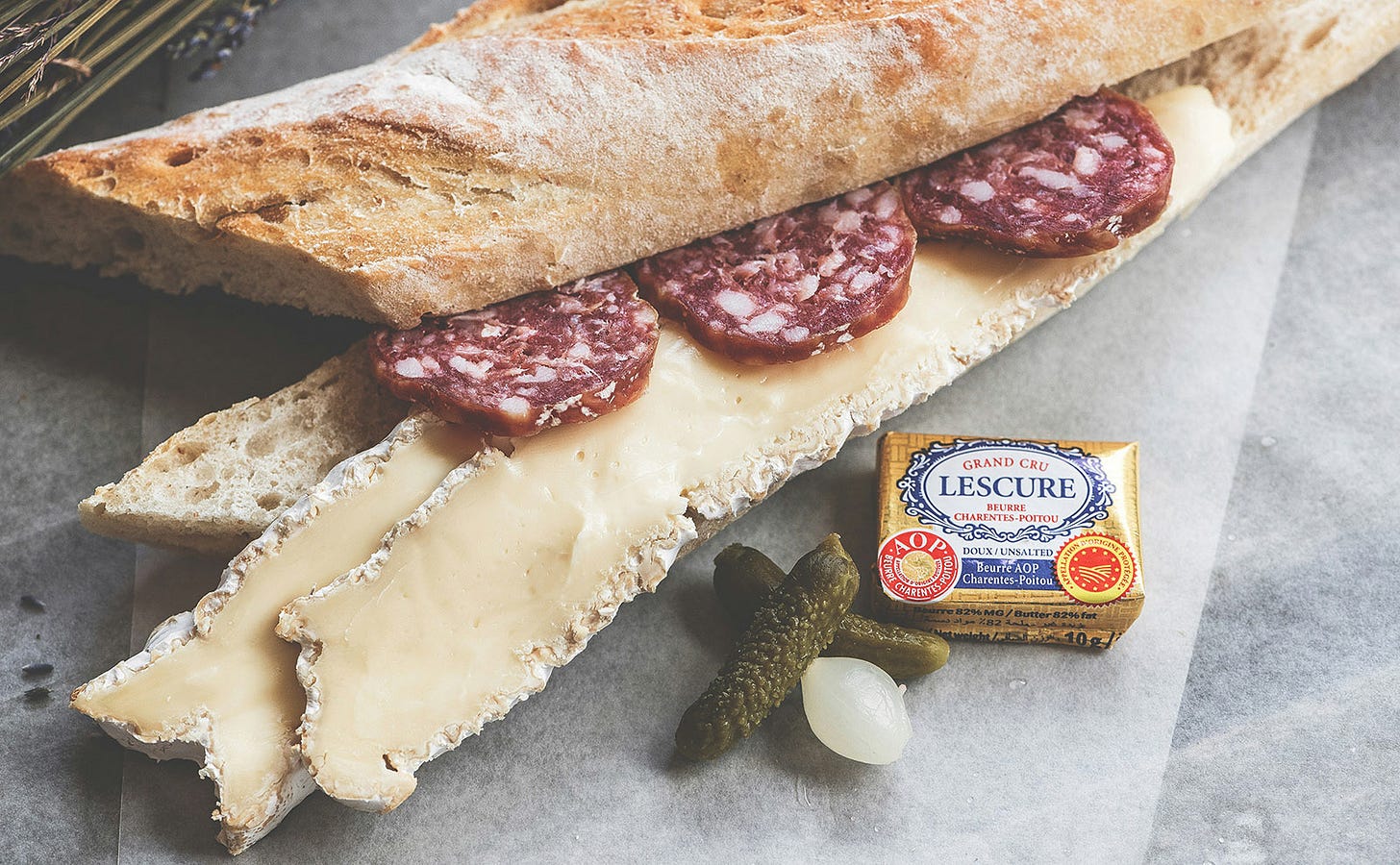 a sliced french baguette on a gray surface with slices of salami and cheese and a pat of butter