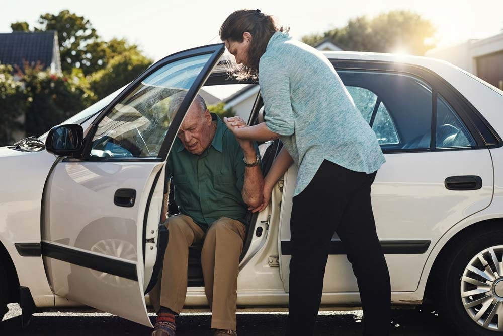 Adult daughter helping her elderly father out of his car because he can’t get out on his own.