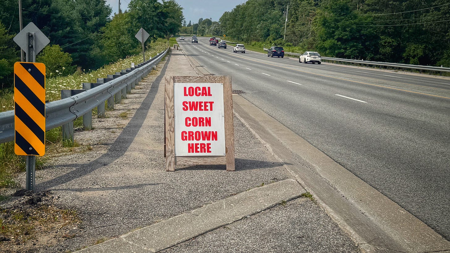 A sign on the shoulder of a busy 4-lane road reads, "LOCAL SWEET CORN GROWN HERE." A sign on the shoulder of a busy 4-lane road reads, "LOCAL SWEET CORN GROWN HERE."