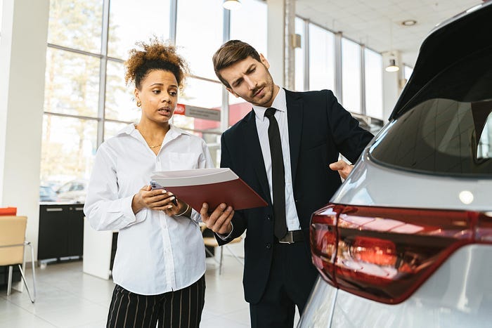 A woman looking concerned while a used car salesman holds up paperwork and is pointing at the car A woman looking concerned while a used car salesman holds up paperwork and is pointing at the car