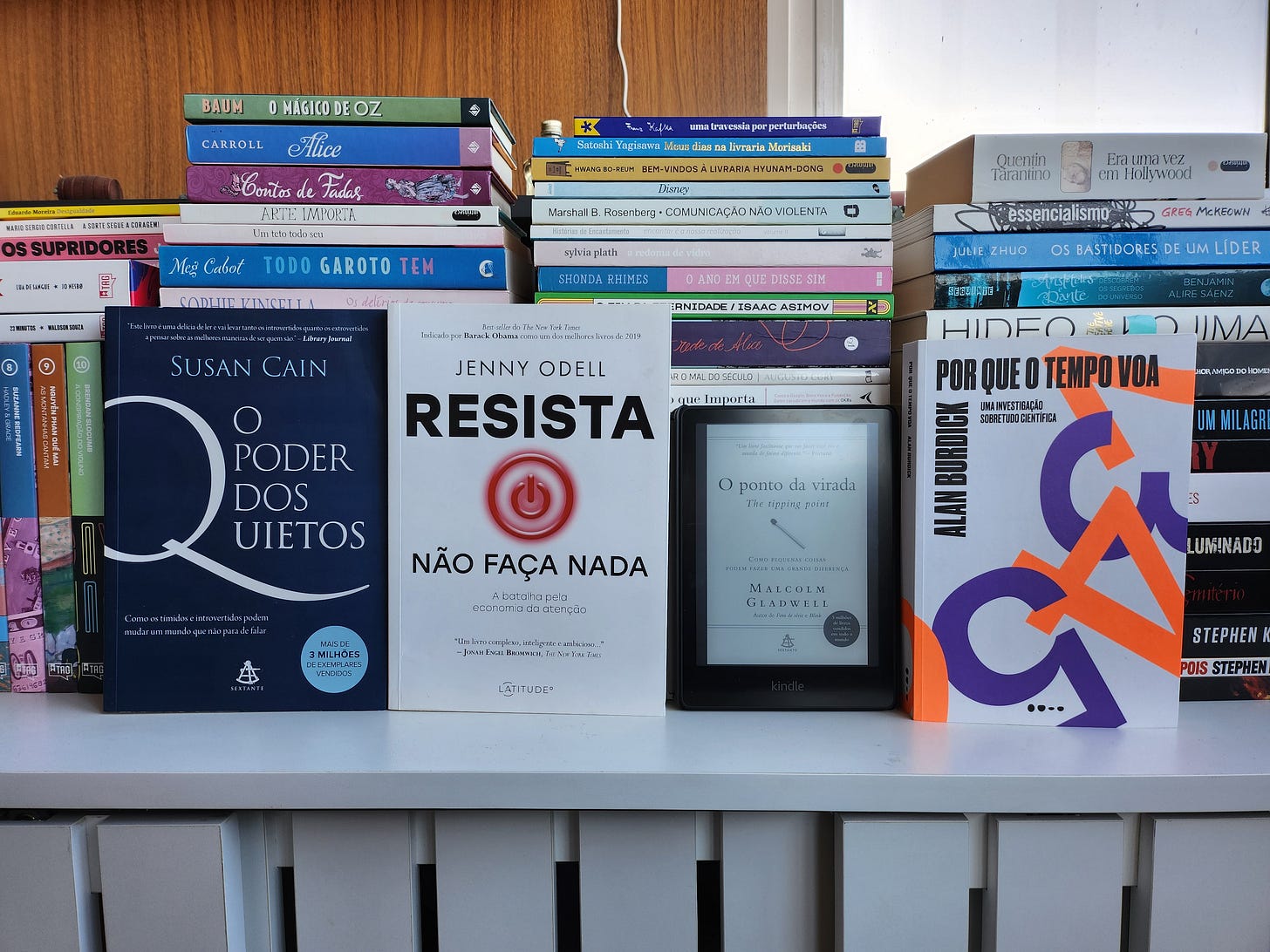 Foto de uma estante com vários livros empinados na horizontal. À frente deles, estão os 4 livros descritos no texto, sendo 3 em edição física e 1 no Kindle.