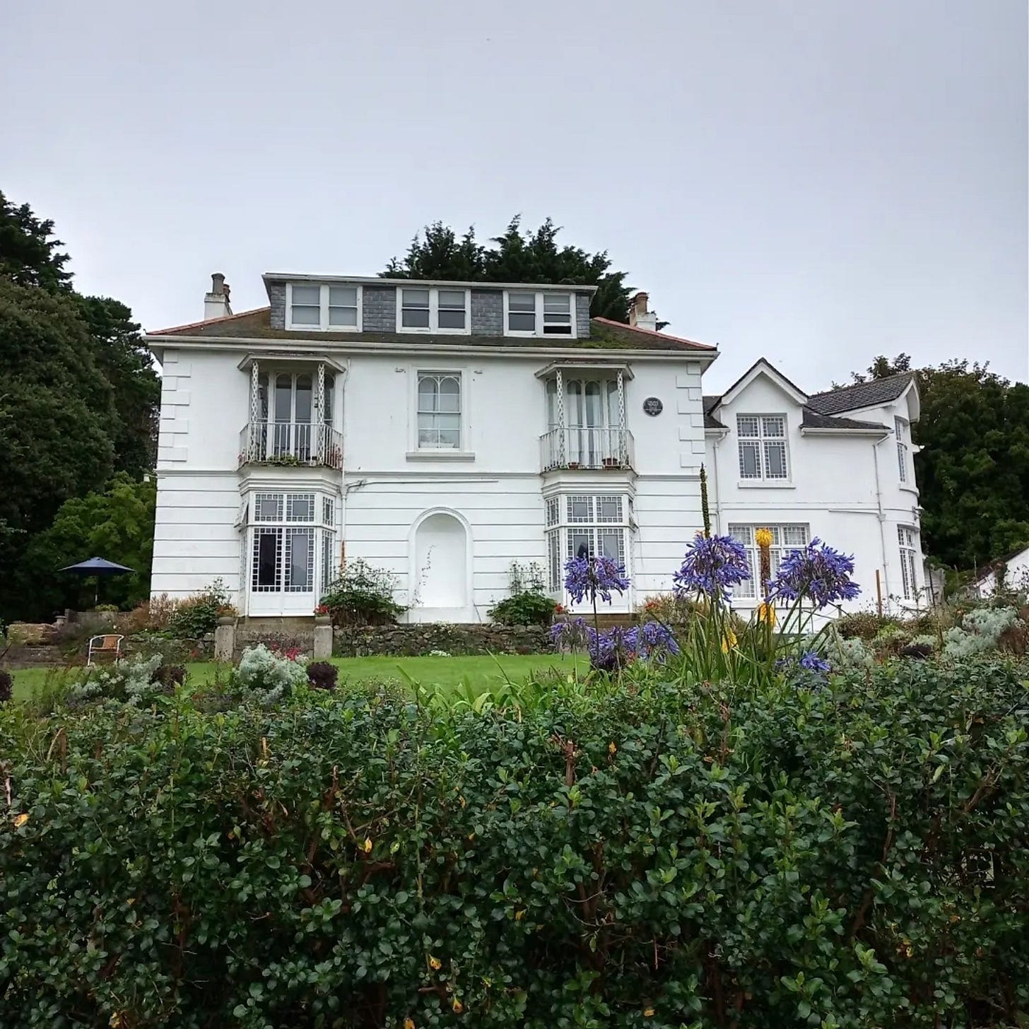 White nineteenth-century two-storey house with large casement windows and dormer roof, above a terrace, lawn and hedge
