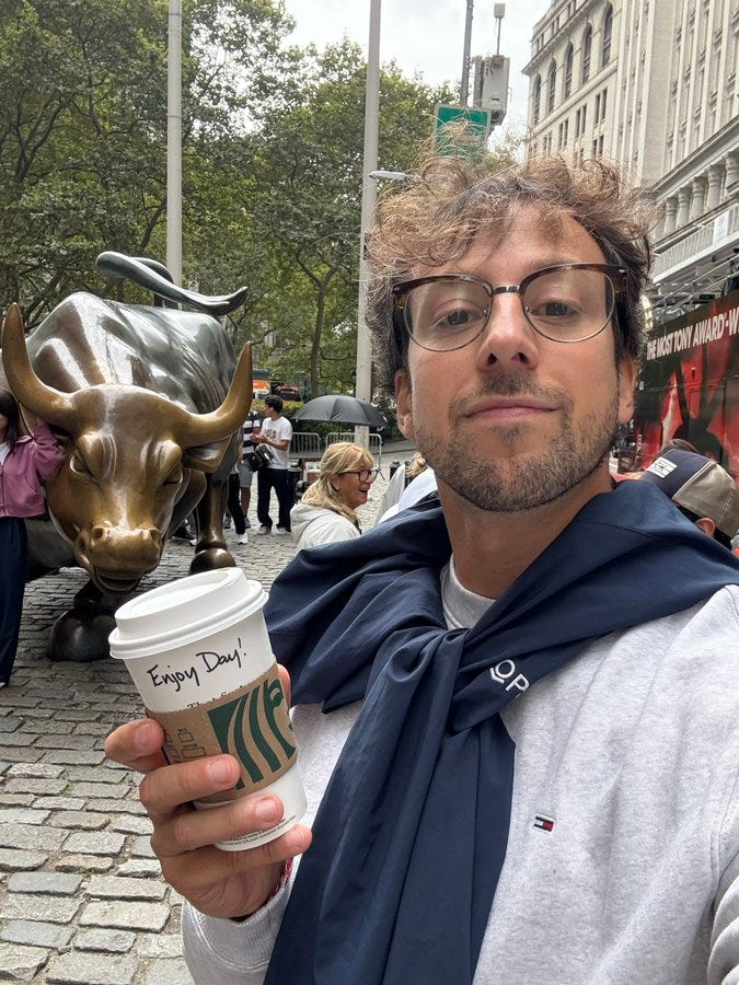 A man with curly hair and glasses holding a coffee cup with the text "Enjoy Day!" on it, wearing a gray hoodie and a dark blue scarf. The Charging Bull statue is visible in the background, set on a cobblestone street with trees and buildings. People are walking nearby. A man with curly hair and glasses holding a coffee cup with the text "Enjoy Day!" on it, wearing a gray hoodie and a dark blue scarf. The Charging Bull statue is visible in the background, set on a cobblestone street with trees and buildings. People are walking nearby.
