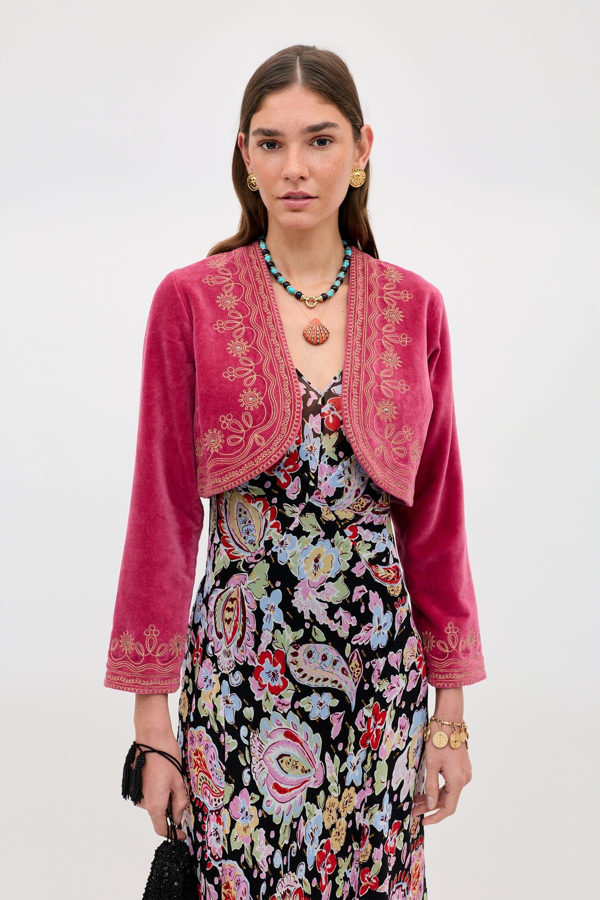 A woman wears the Cliona - Soutache Embroidery Magenta Pink jacket over a floral dress, accessorized with gold jewelry and a black bag, standing against a plain light background.