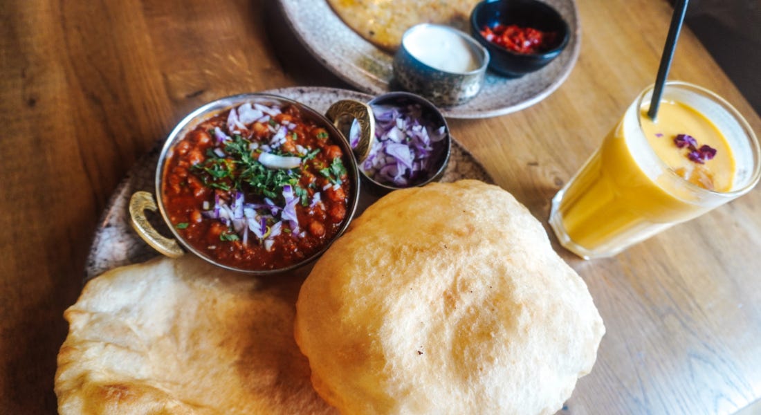 A wooden table with two plates of Indian food with bowls and curries, with a mango lassi glass