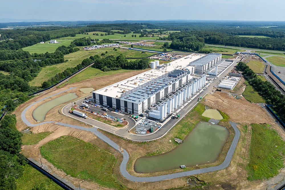 Aerial view of a data center - a large, white rectangular building with several cooling tanks and generators jutting out along the side and on the roof - with runoff water drainage on either side of one end. The data center is surrounded by green trees, fields and farmland. Aerial view of a data center - a large, white rectangular building with several cooling tanks and generators jutting out along the side and on the roof - with runoff water drainage on either side of one end. The data center is surrounded by green trees, fields and farmland.