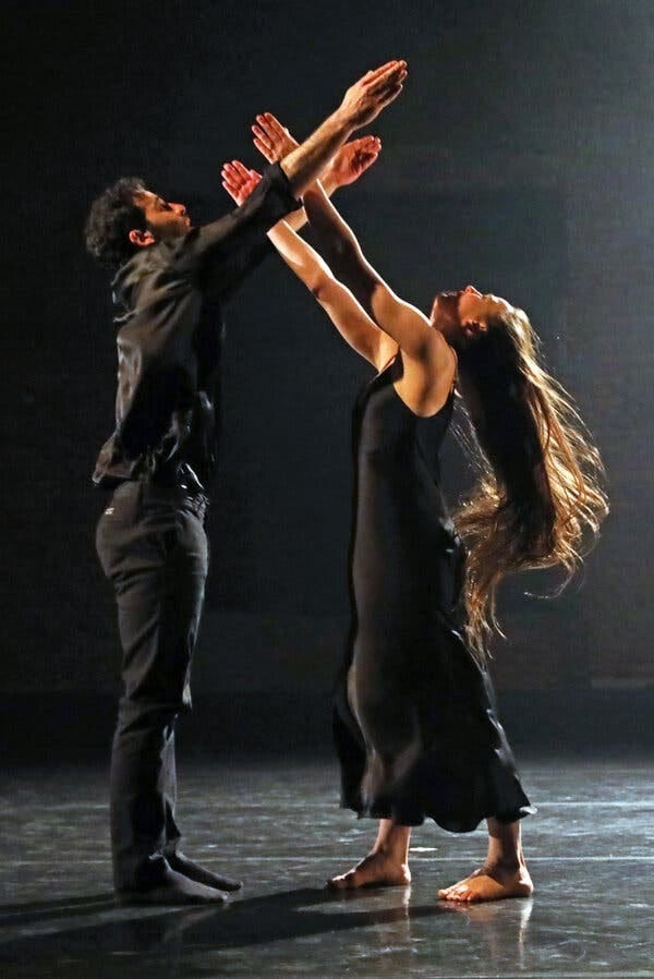 A man and woman are on a stage, both dressed in black. Their arms are outstretched upward and they are looking up.