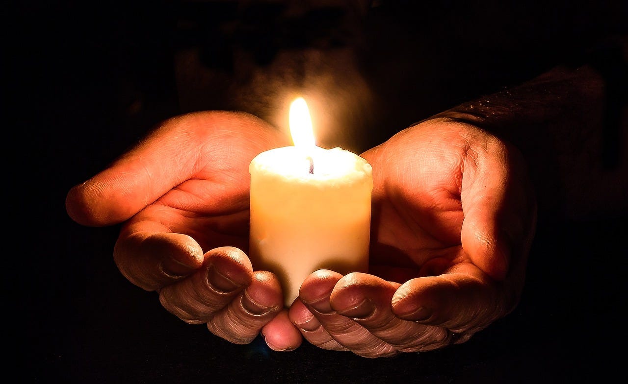 Black background, two hands holding a lit yahrzeit candle