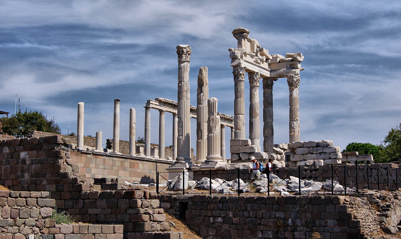 A picture of the ancient ruins of Pergamum on a blue sky day. The broken marble pillars reach into the sky to where a roof would have once been. Tourist walk around the ruins in the forground. A picture of the ancient ruins of Pergamum on a blue sky day. The broken marble pillars reach into the sky to where a roof would have once been. Tourist walk around the ruins in the forground.