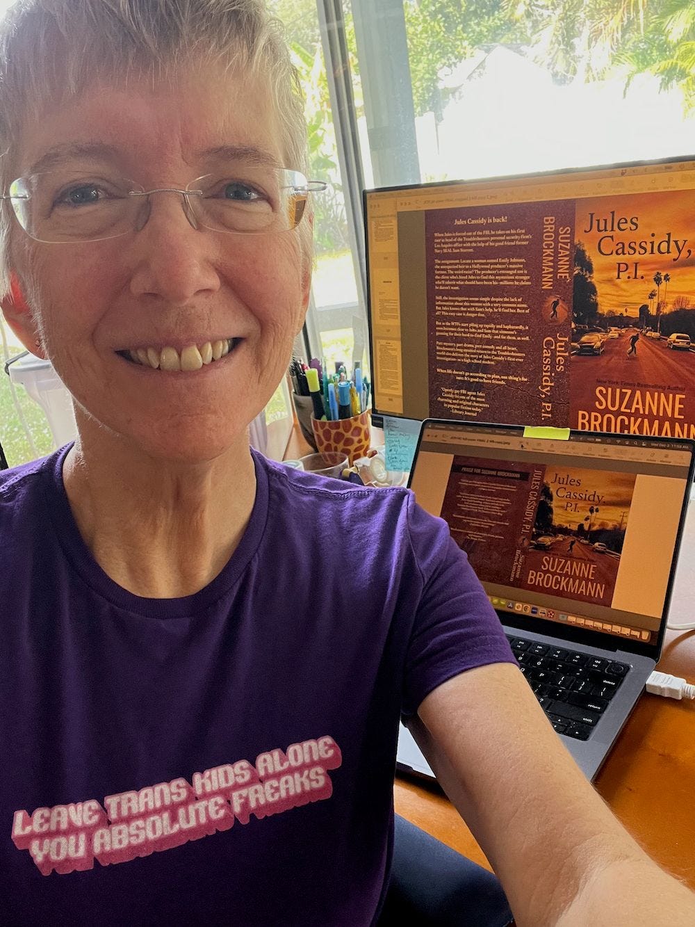 Your author, Suz Brockmann, a tired-looking older white woman with short fair hair and glasses wears a purple T-shirt reading “Leave trans kids alone you absolute freaks.” She smiles as she takes a selfie in front of her computer screens, which hold the print editions’ cover art to her latest book, Jules Cassidy, P.I. The cover art shows a reddish-tinged evening sky and a road in a tired-looking Los Angeles Valley neighborhood where the small figures of two men, one large, one smaller, run across the street. Palm trees and electrical poles are silhouette against the sunset.