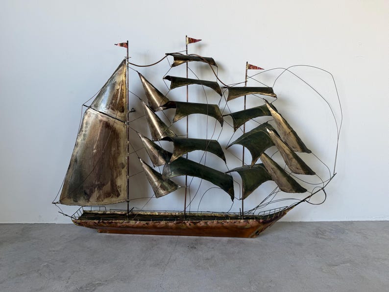 May include: A metal sailboat sculpture with a brown hull, silver sails, and green and gold accents. The sculpture has red flags on the masts and is displayed against a white wall.