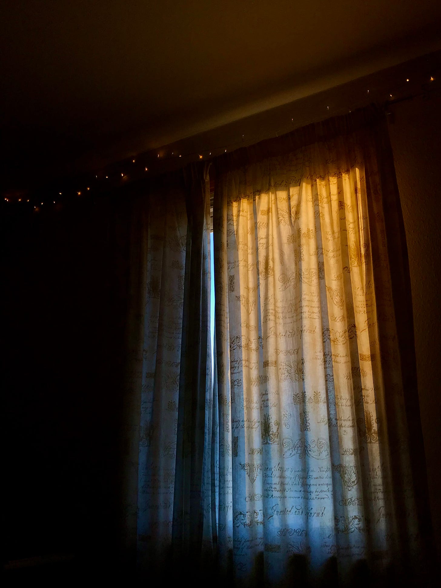 Early morning or late evening sunlight filters gold and indigo through a window covered with curtains. 