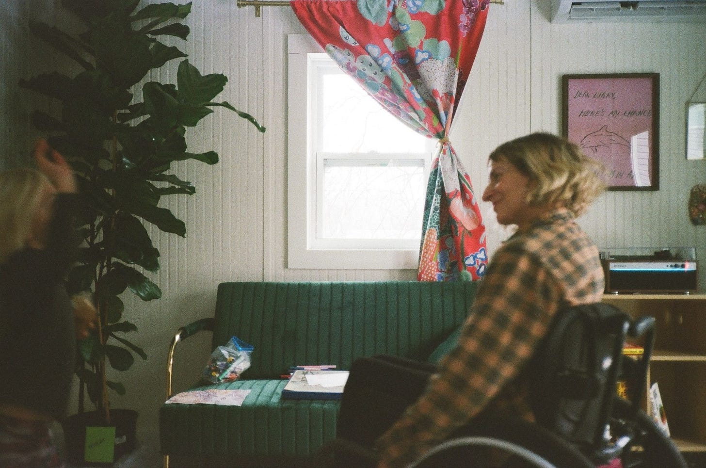 A photo in Rebekah’s office — a plant, a couch, a curtain, a picture on the wall. She sits in her wheelchair, grinning at her son — a blur on the left edge of the photo, one hand up. 