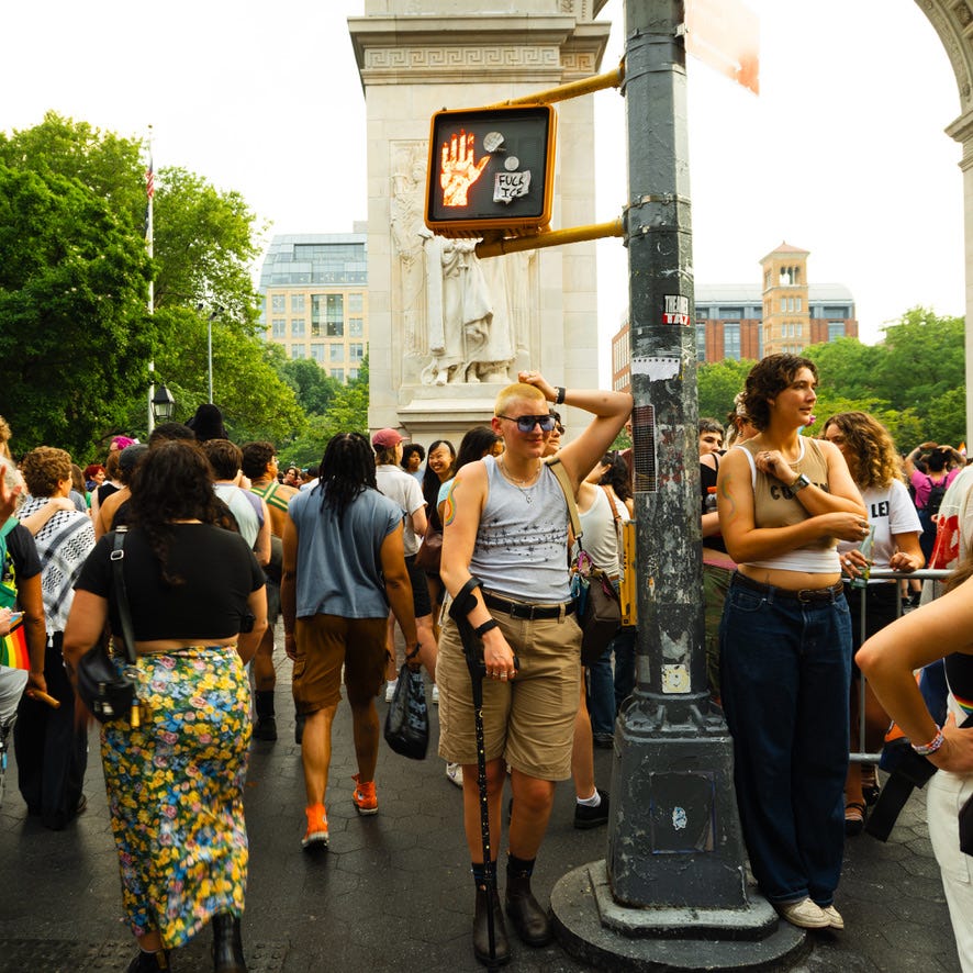 The photographer leaning against a traffic pole, slightly smiling and holding their mobility aid.