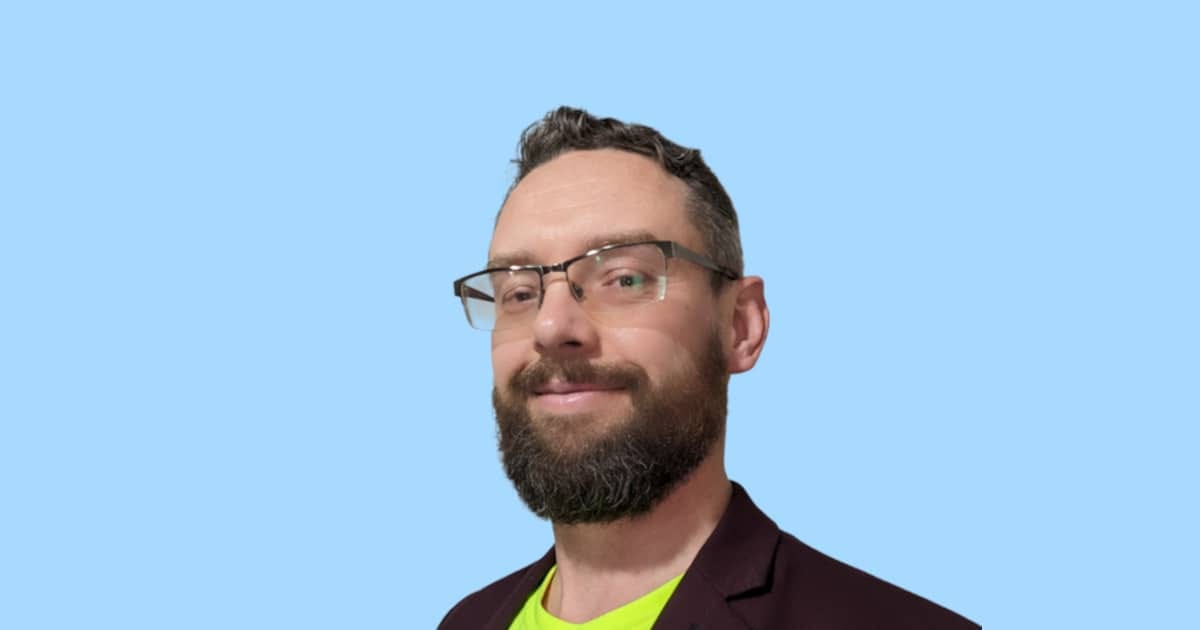 A close-up studio portrait of Roundism founder Michal Plis, a bearded man wearing glasses and a dark jacket against a solid light blue background. A close-up studio portrait of Roundism founder Michal Plis, a bearded man wearing glasses and a dark jacket against a solid light blue background.
