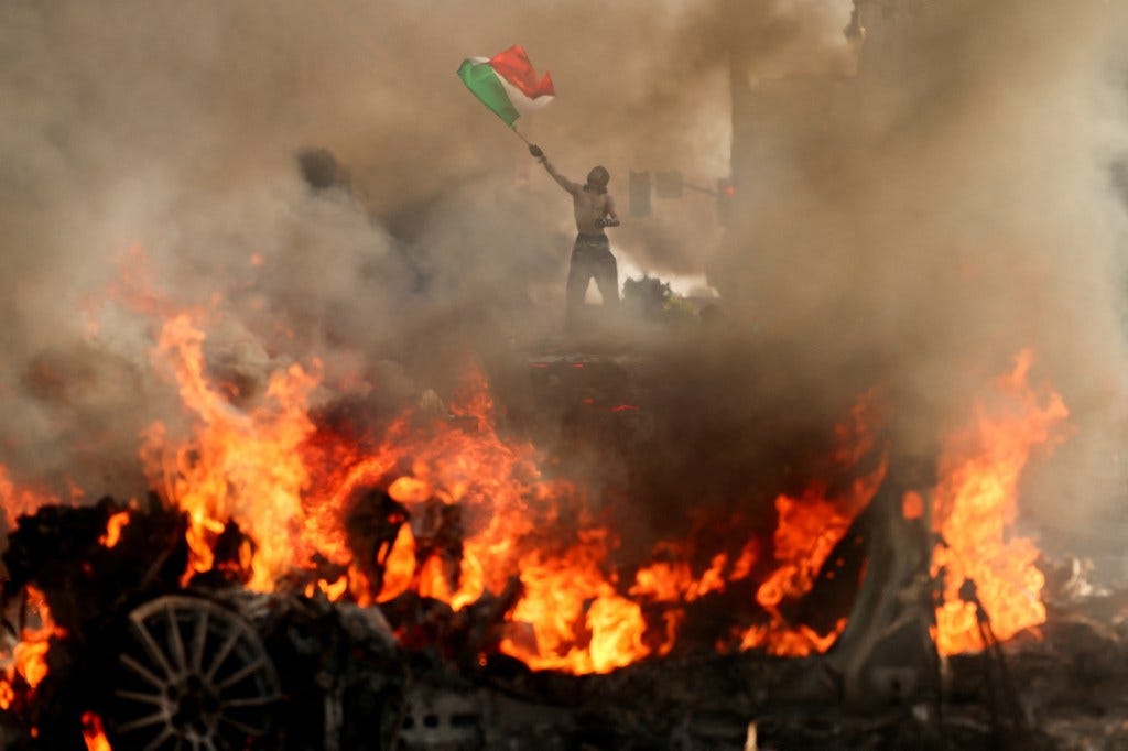 A man waves a Mexican flag as smoke and flames rise from a burning vehicle