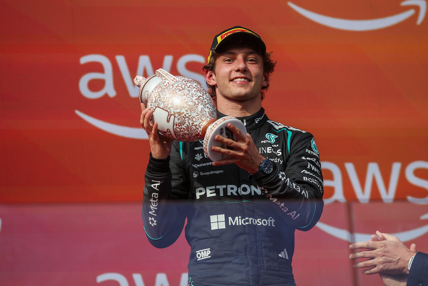 Kimi Antonelli stood on an F1 podium wearing black Mercedes race overalls and smiling while holding a porcelain trophy