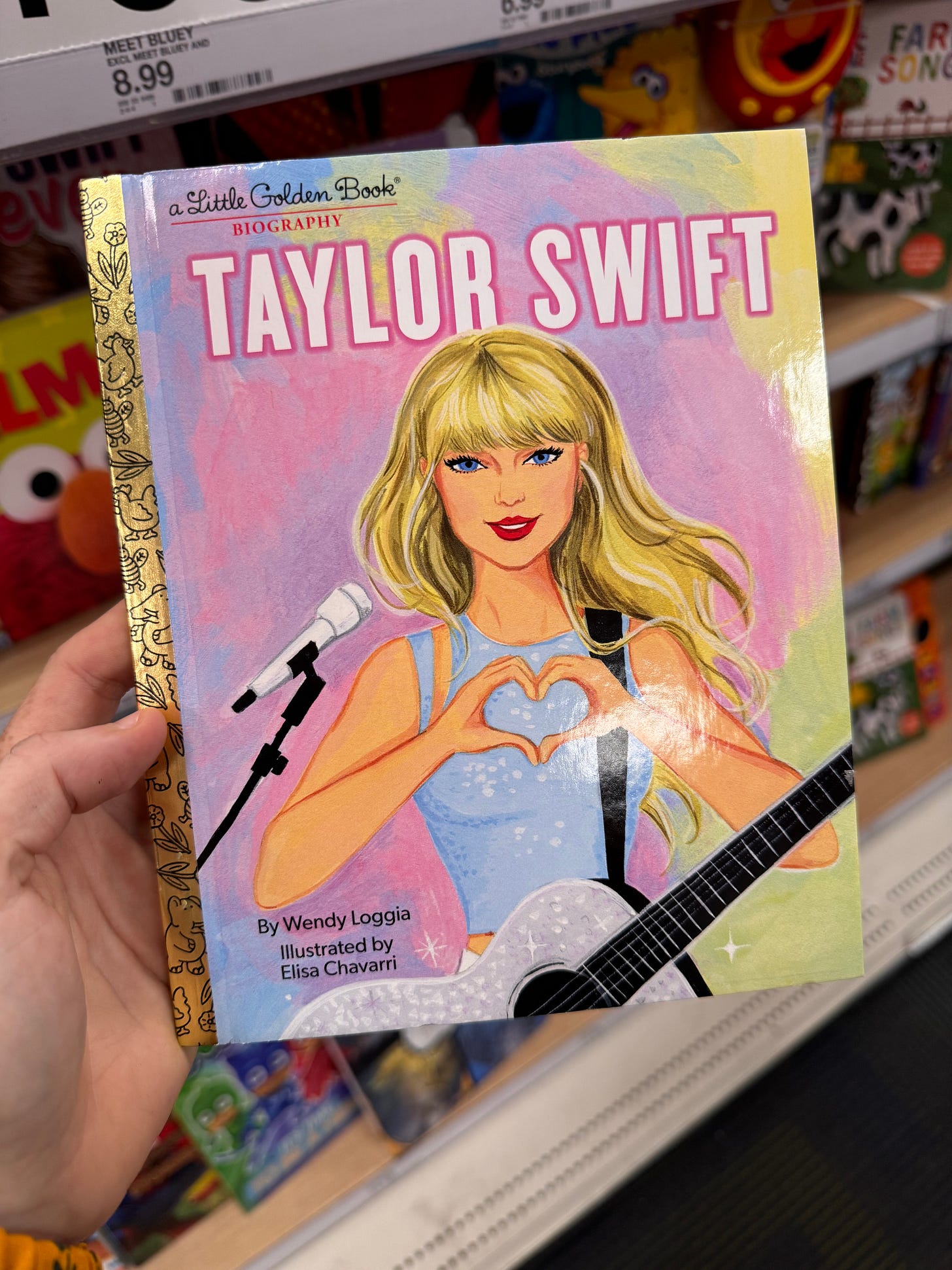 Cover of "Little Golden Book Biography" of Taylor Swift. Children's book with a golden spine and an illustrated image of Taylor making a heart shape with her hands, as pastel rainbow colors sparkle in the background.
