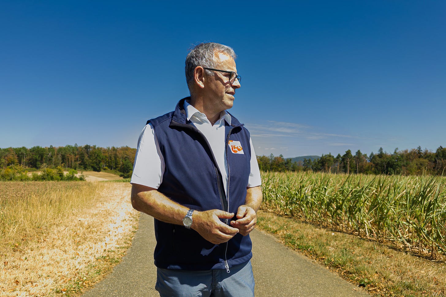 Christoph Böbner, Leiter des Ebenrain-Zentrums, steht bei sommerlicher Hitze auf einem Feldweg zwischen einem ausgetrockneten Feld und einem Maisacker. Im Hintergrund sind trockene Vegetation und Waldrand unter wolkenlosem blauem Himmel zu sehen.